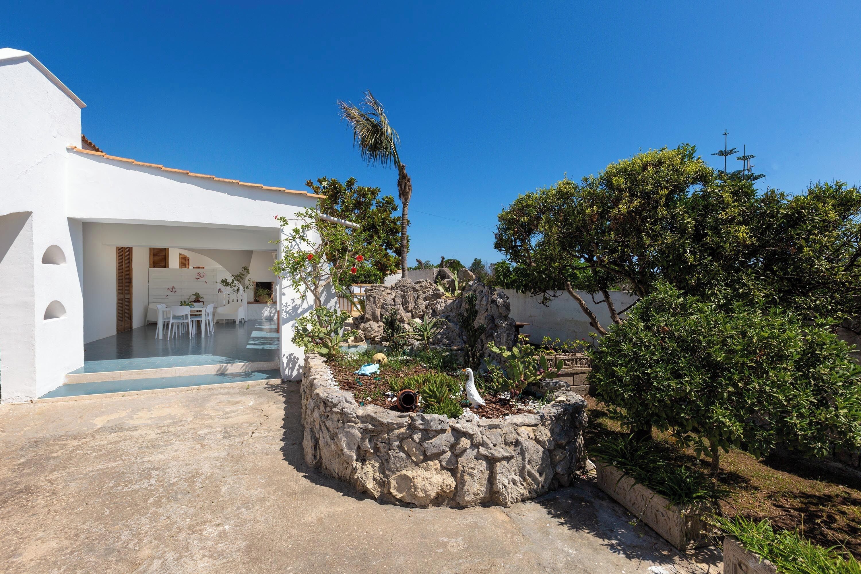 A white building with a stone garden, a palm tree, and a clear blue sky.