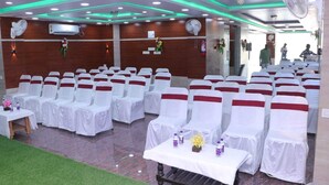 A banquet hall with rows of white chairs with red sashes, tables with white tablecloths, and green ambient lighting. - Royal Park Celebration By GRB (Patna)