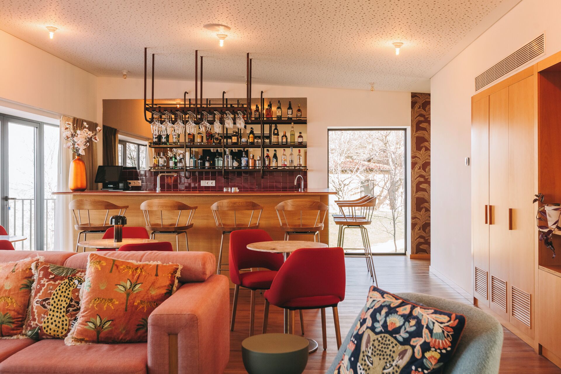 Uma área de bar moderna com diversos frascos de bebidas, um espaço de estar com cadeiras vermelhas e uma grande janela com vista para o exterior.
