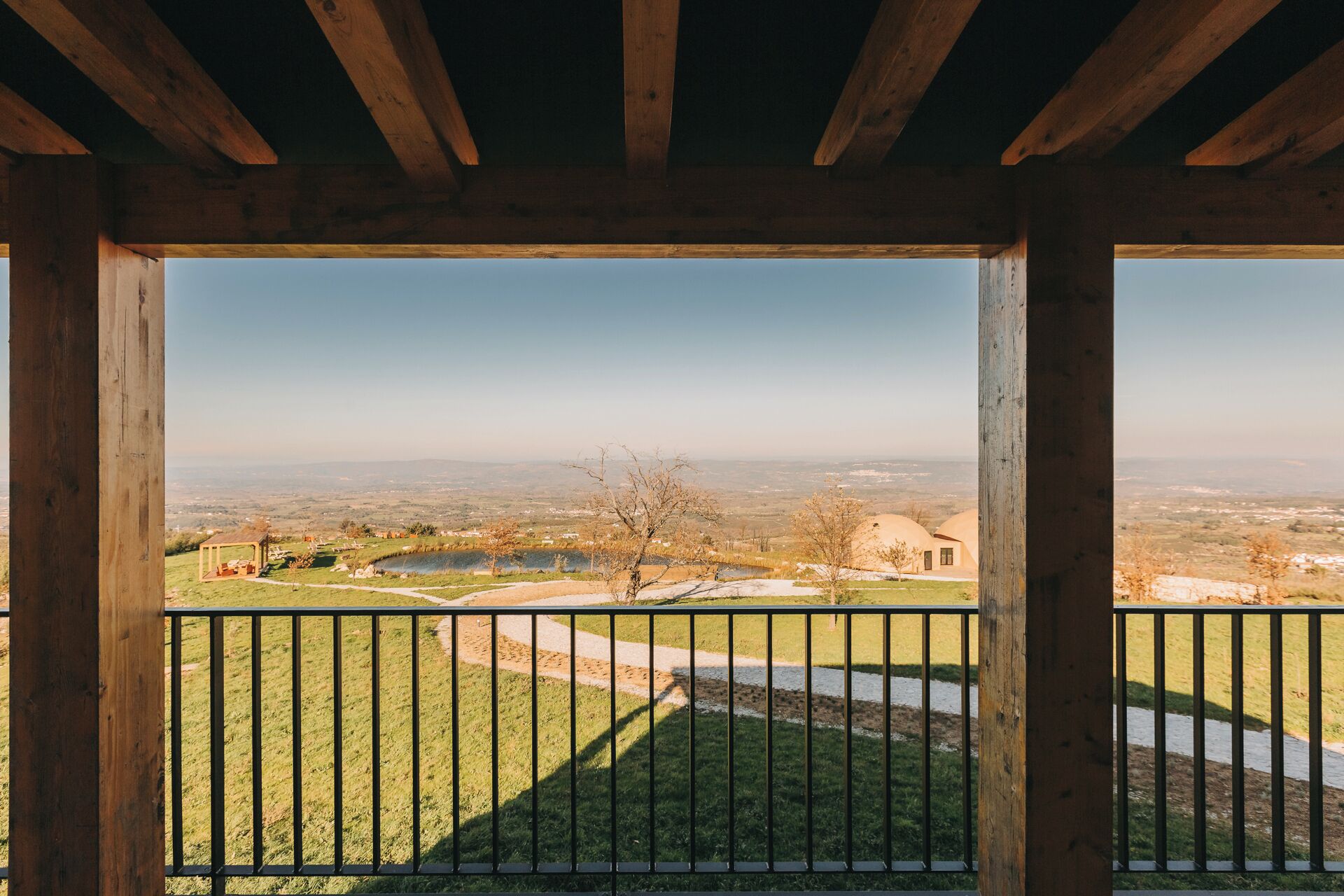 A wooden balcony with a view of a rural landscape, including a pond, trees, and distant buildings.