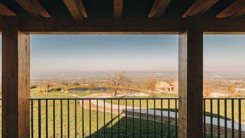 A wooden balcony with a view of a rural landscape, including a pond, trees, and distant buildings.