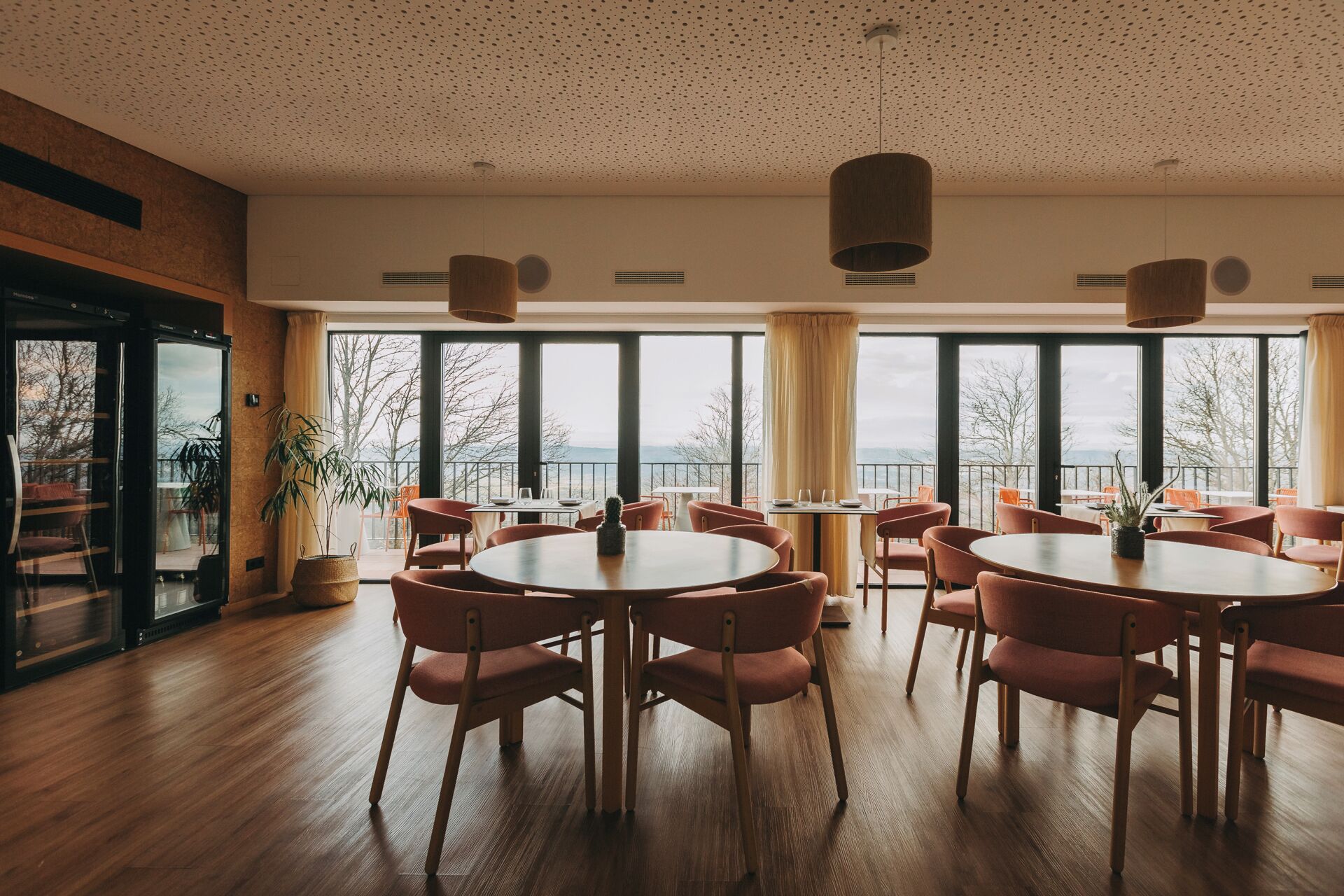 Un vaste espace dédié à la salle à manger, avec des tables et des chaises rondes, de grandes fenêtres offrant une vue sur l’extérieur et un plafond orné d’un motif moucheté.