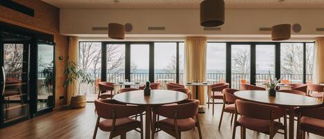 A spacious dining area with round tables and chairs, large windows offering a view of the outdoors, and a ceiling with a speckled pattern.