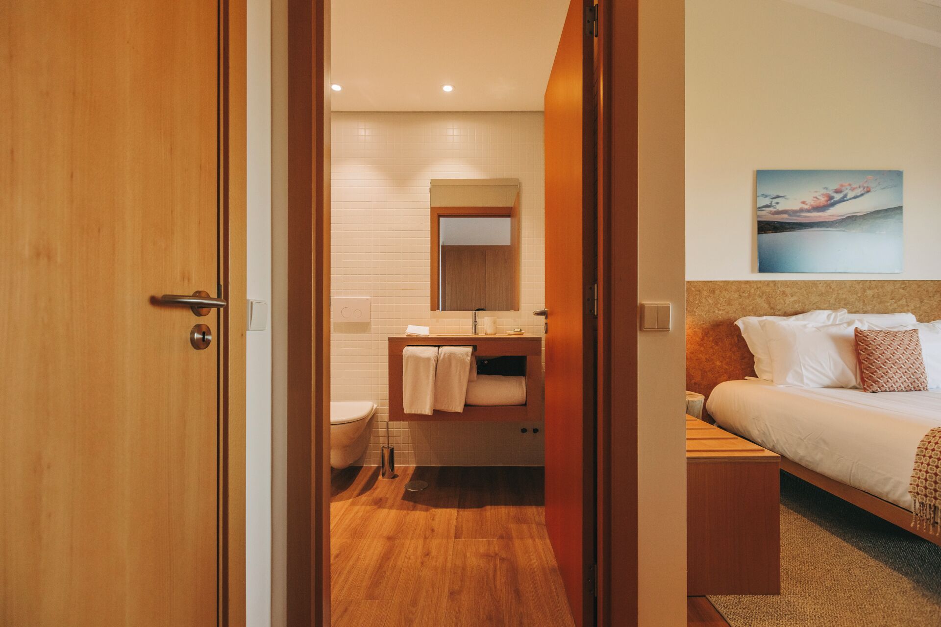 Een hotelkamer met een houten deur, een badkamer met toilet, wastafel en spiegel, en een bed met wit beddengoed en een schilderij erboven.