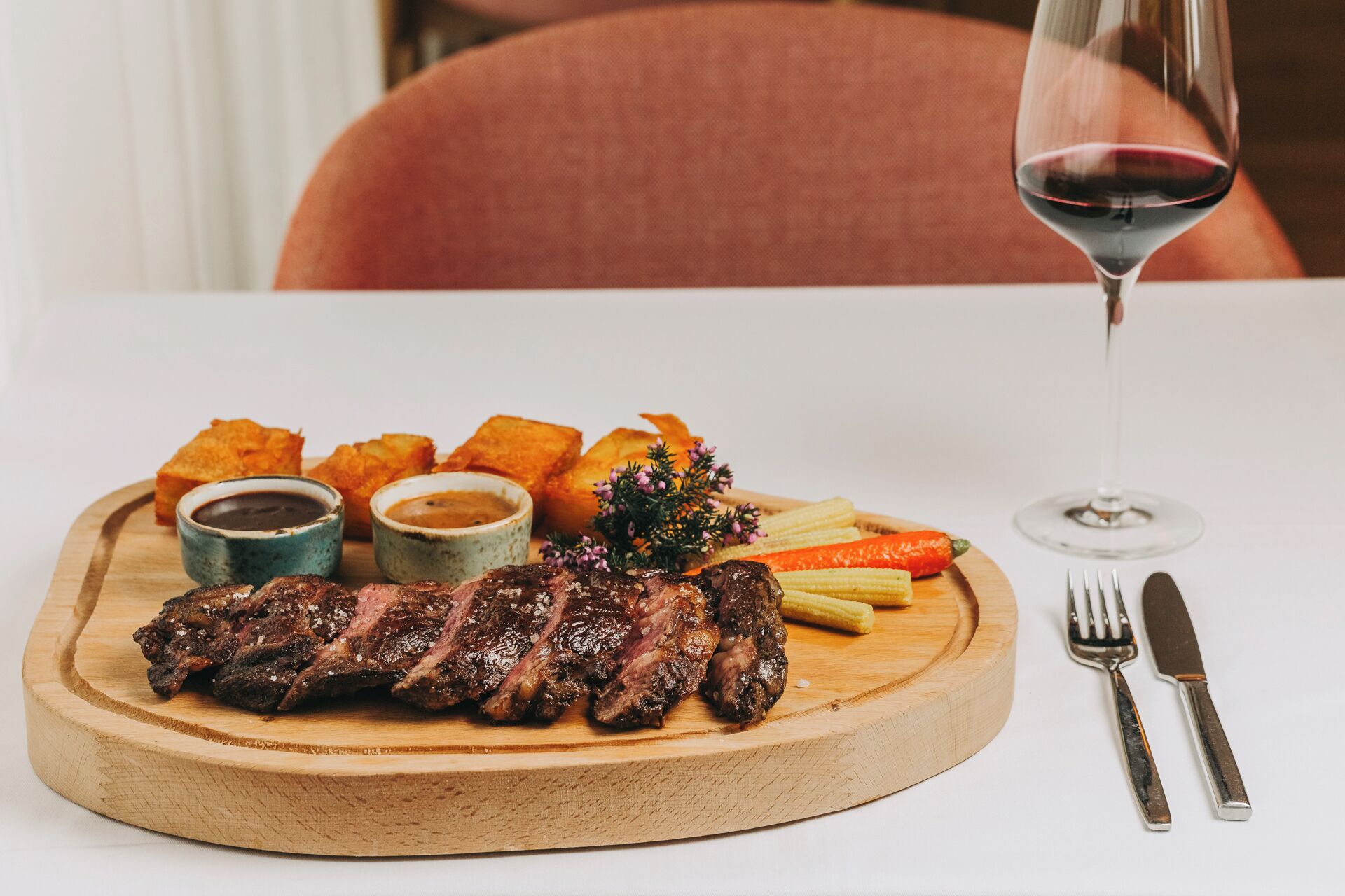 A wooden board with grilled meat, fries, and two dipping sauces, accompanied by a glass of red wine and cutlery on a white table.
