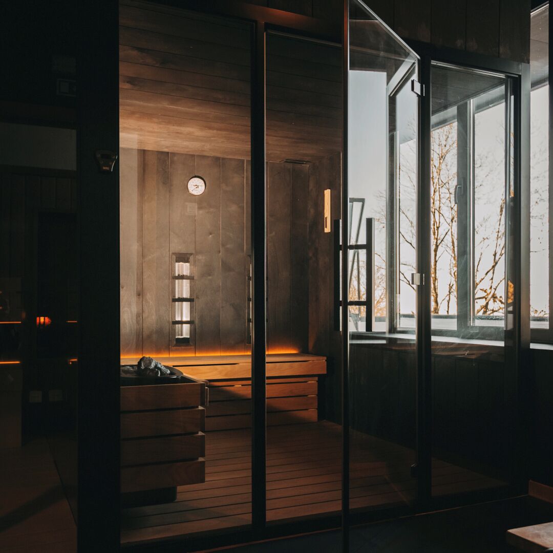 Un intérieur moderne avec un plancher en bois, un banc, une horloge murale et une grande fenêtre donnant sur des arbres sans feuilles.