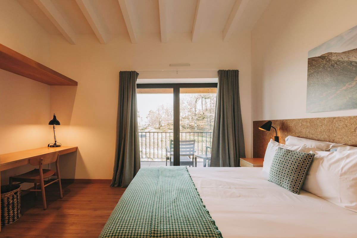 Et hotelværelse med seng, skrivebord, stol, en balkon med gardiner og et billede på væggen.