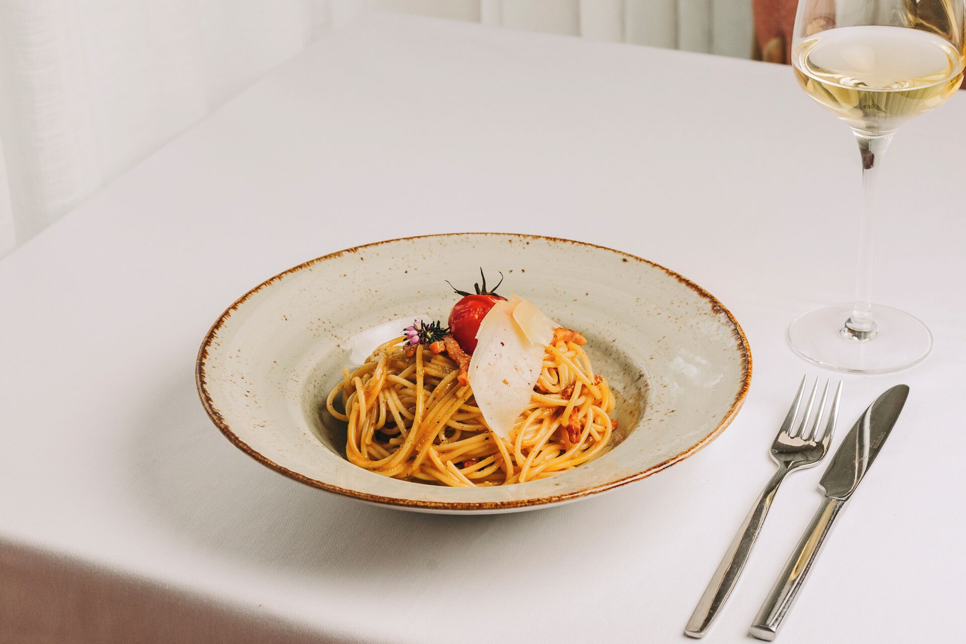Une assiette de spaghettis garnie de tomates cerises et d’une tranche de fromage, le tout présenté sur une assiette à motifs, accompagné d’un verre de vin blanc et de couverts posés sur une nappe blanche.