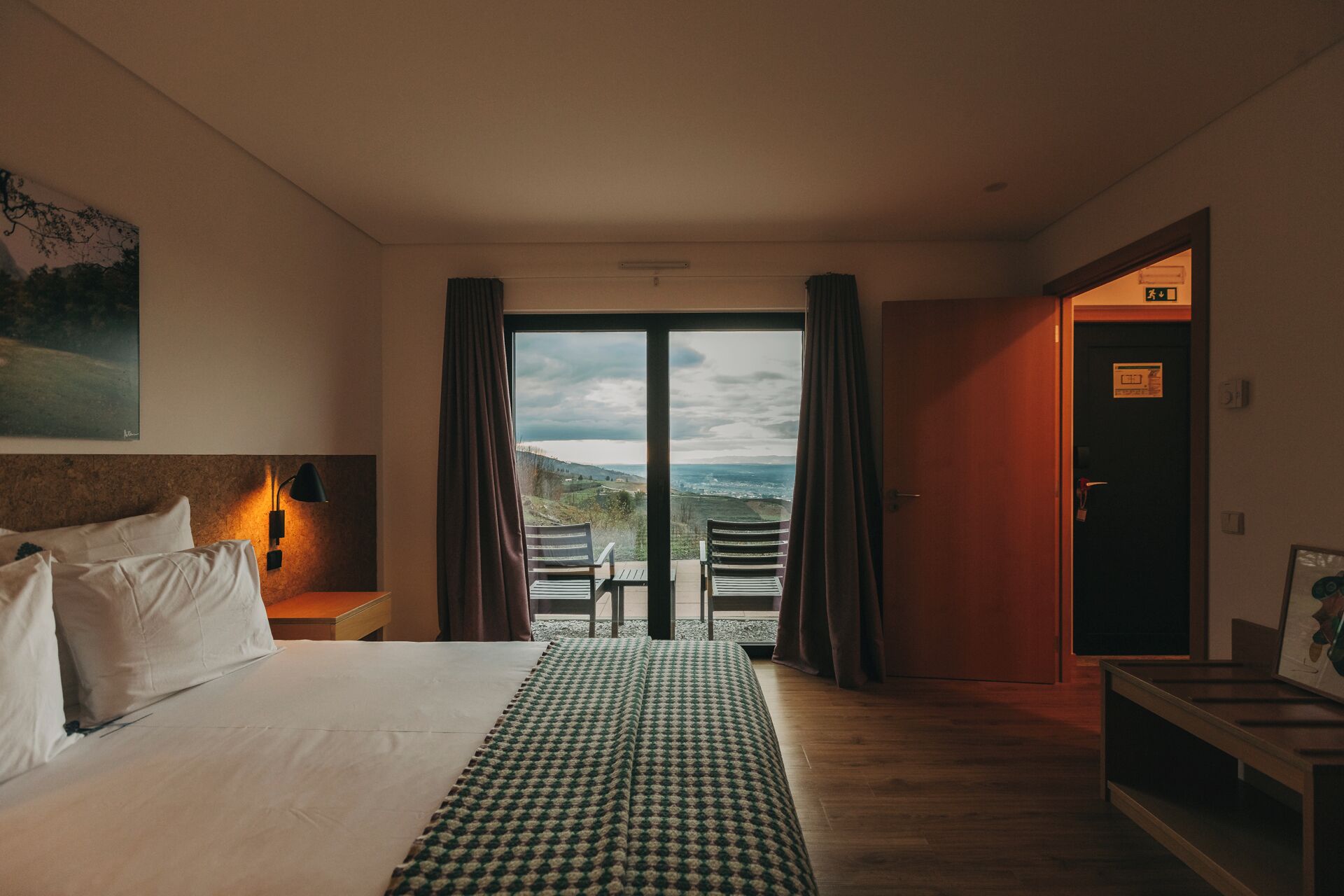 A hotel room with a large bed, a desk with a lamp, a view of the countryside through the window, and a door with a sign.