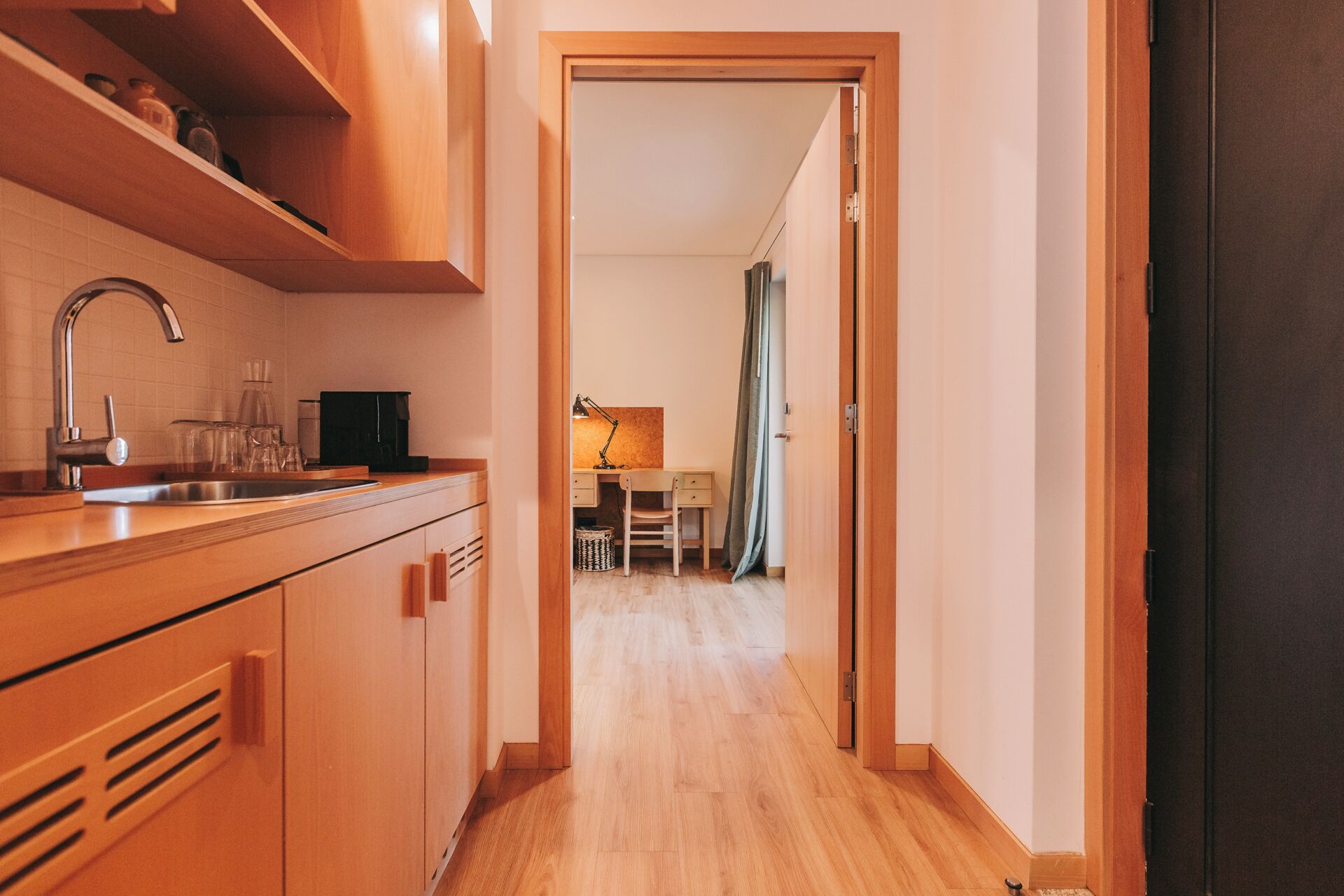 Un estrecho pasillo con armarios de madera y un lavabo a la izquierda. Se ve un escritorio con una silla y una lámpara al fondo.