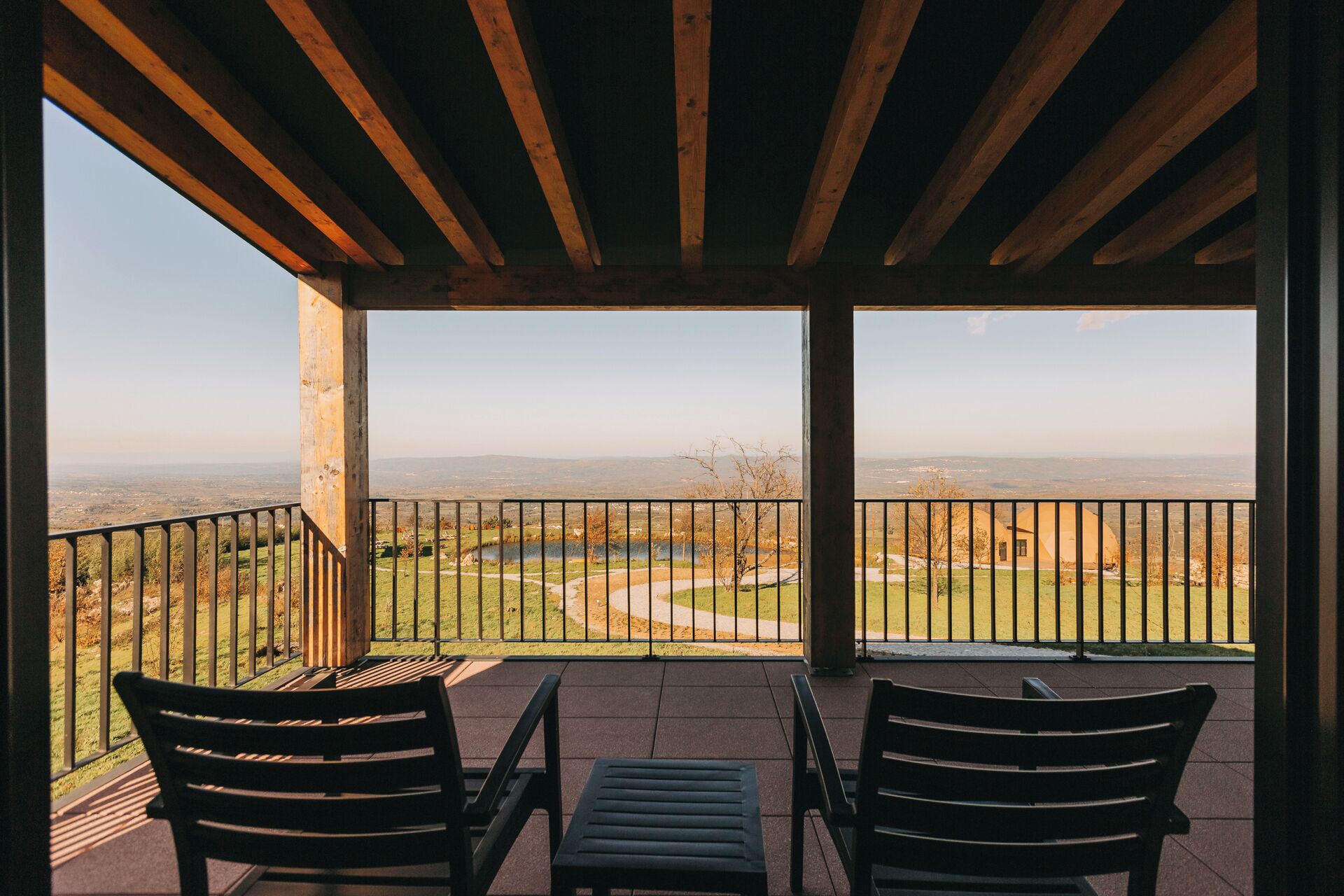 Een overdekt terras met twee stoelen en een tafel, met uitzicht op een landschap met een waterpartij en heuvels in de verte.