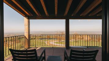 Uma área de estar coberta ao ar livre com duas cadeiras e uma mesa, com vista para uma paisagem com um corpo d'água e colinas ao longe.