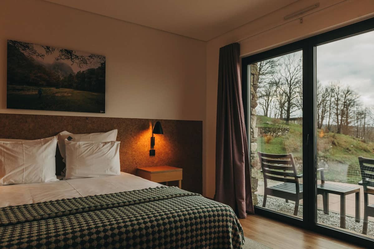 Et hotelværelse med en stor seng, et natbord, en vægmonteret lampe, et billede og udsigt til det fri gennem en skydedør.