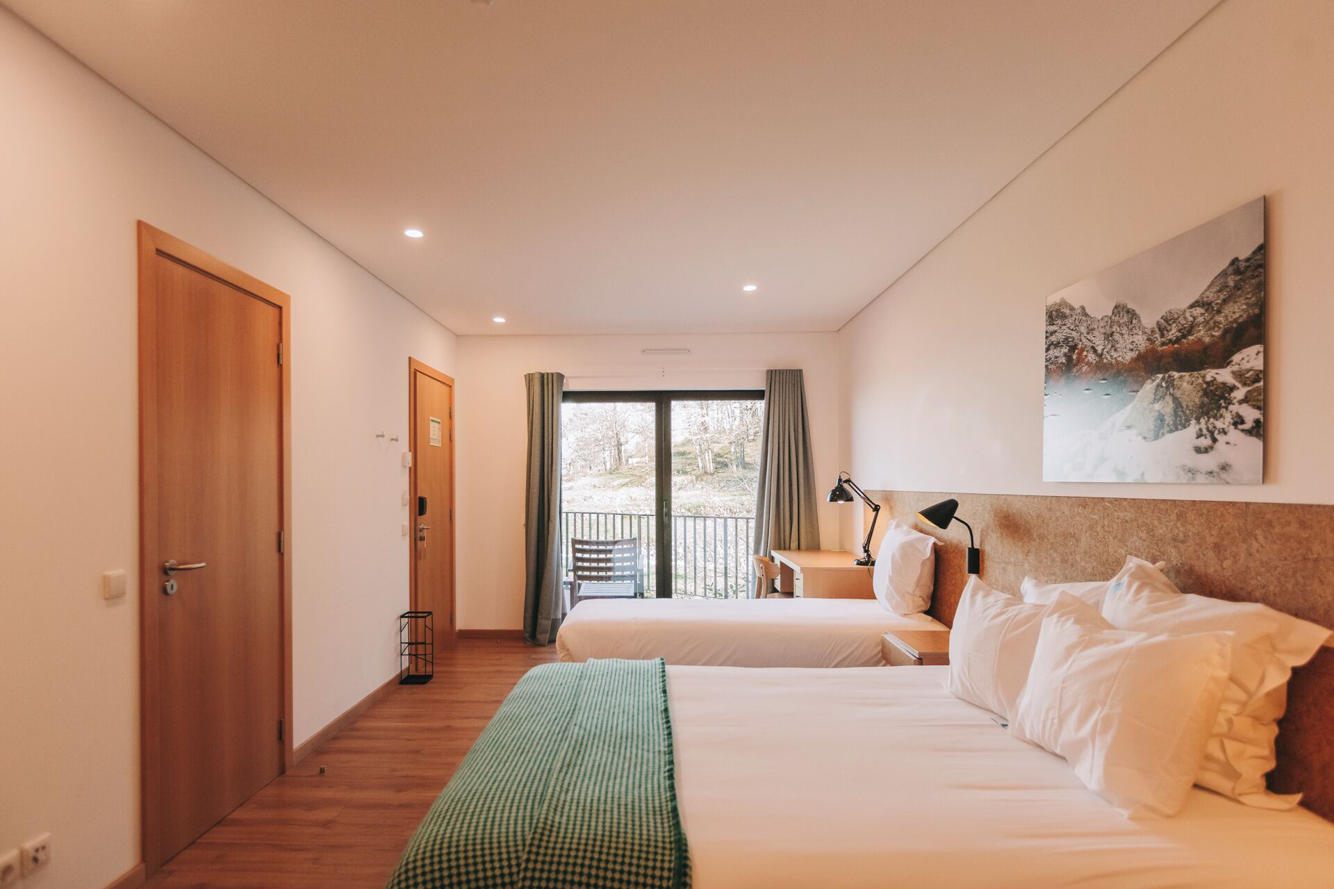 Habitación de hotel con dos camas, un escritorio, una silla, una lámpara y un amplio ventanal con cortinas.