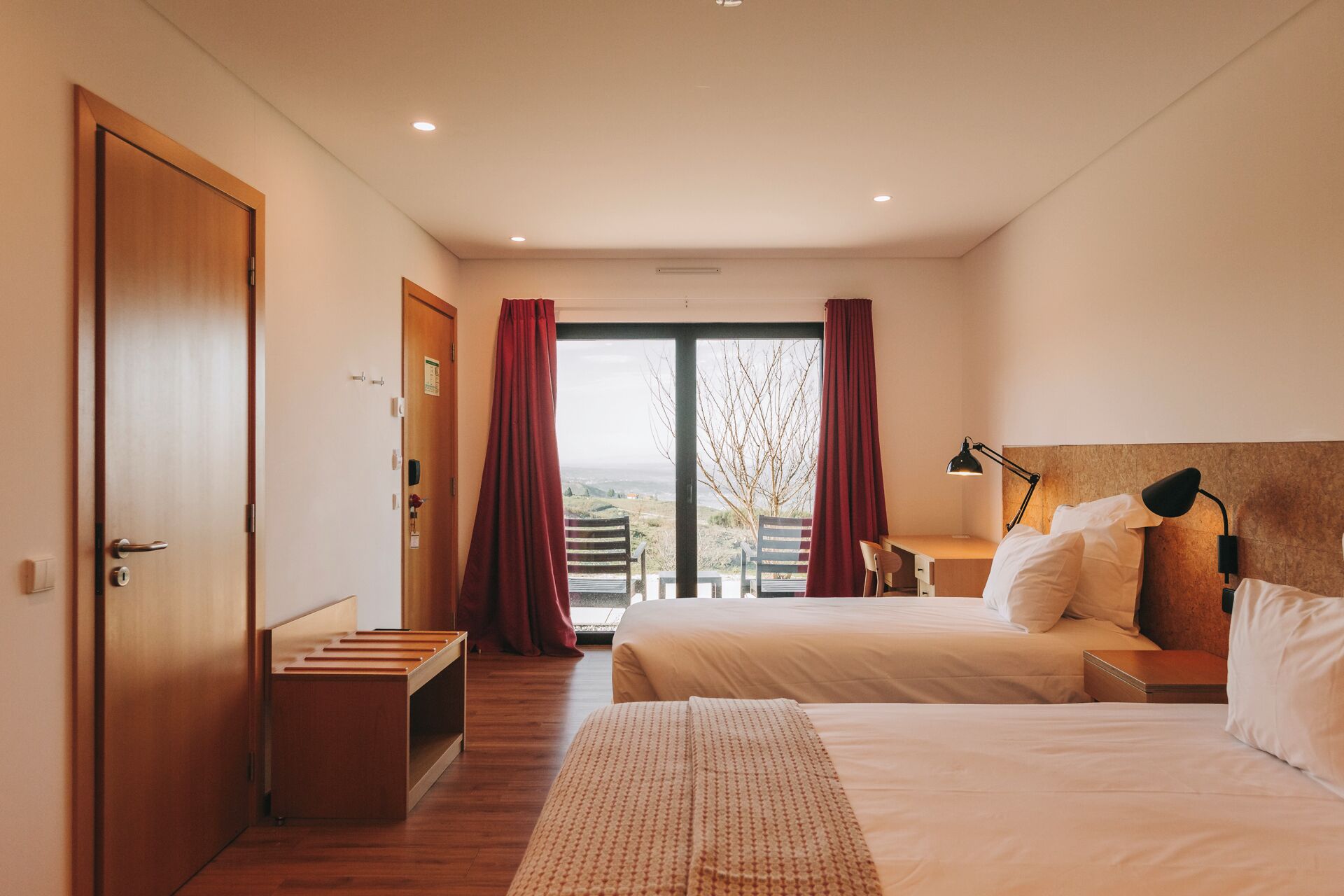 A hotel room with two beds, a desk, a chair, and a view of the outdoors.