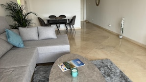 Een moderne woonkamer met een grijze modulaire bank, een ronde salontafel met een tijdschrift, een potplant en een eethoek op de achtergrond.