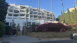 Een hotel met meerdere verdiepingen, balkons, palmbomen en een stenen muur met roze bloemen.