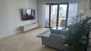 Een woonkamer met een grijze bank, een flatscreen televisie aan de muur, een kleine salontafel en een potplant.