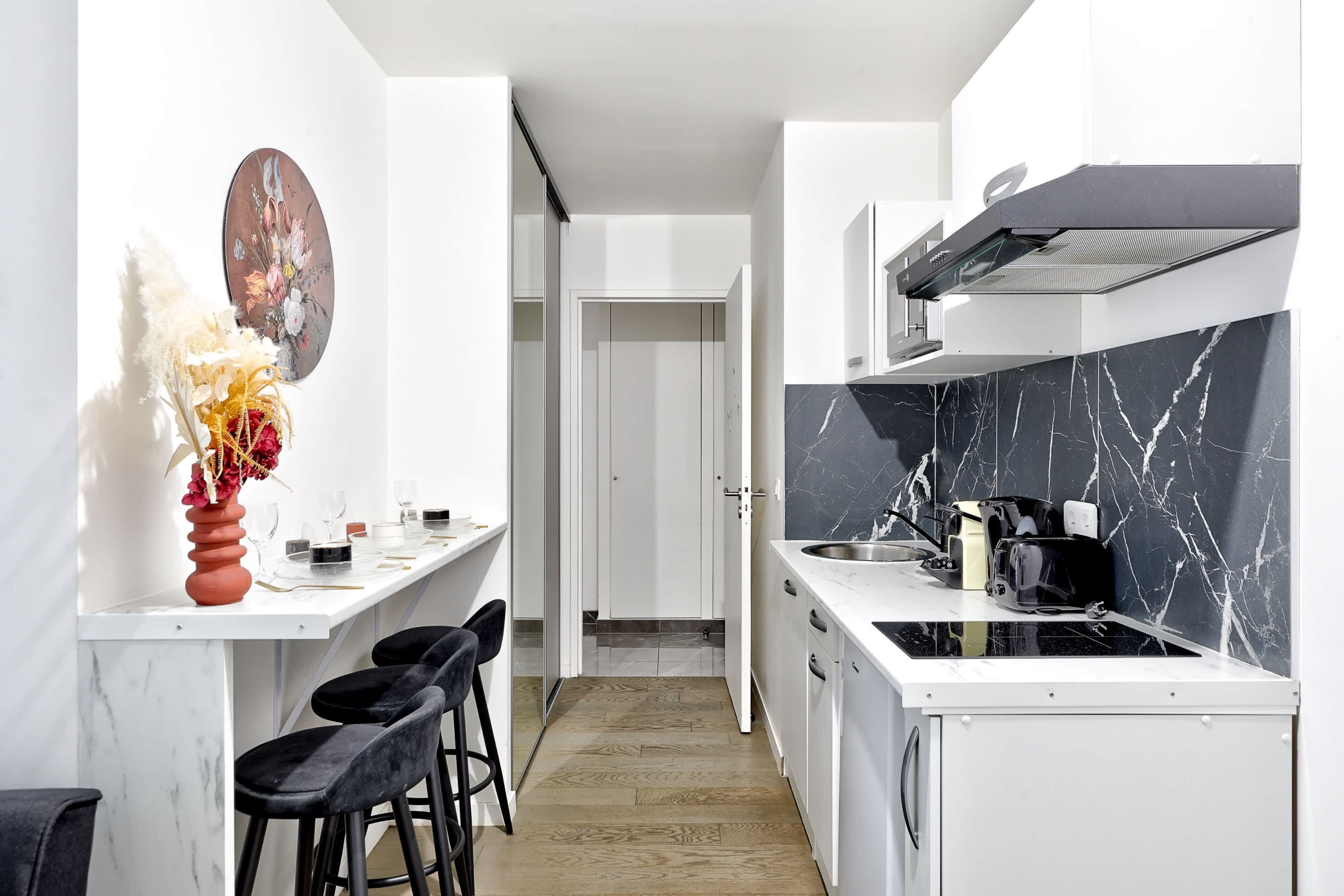 A narrow kitchen with a white countertop, black marble backsplash, and a black stove. There are two black bar stools and a vase with dried flowers on the left.