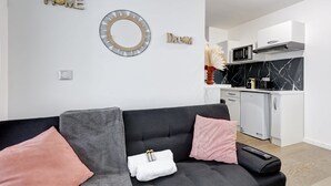 Ein modernes Wohnzimmer mit grauem Sofa, einer kleinen Küche im Hintergrund und dekorativen Wandbildern.
