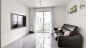 A modern living room with a large flat-screen TV, a black leather sofa, and a sliding glass door leading to a balcony.