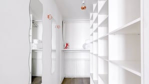 A narrow hallway with white walls and a wooden floor. It features a white radiator, a mirror, and a white shelving unit with a red object on top.