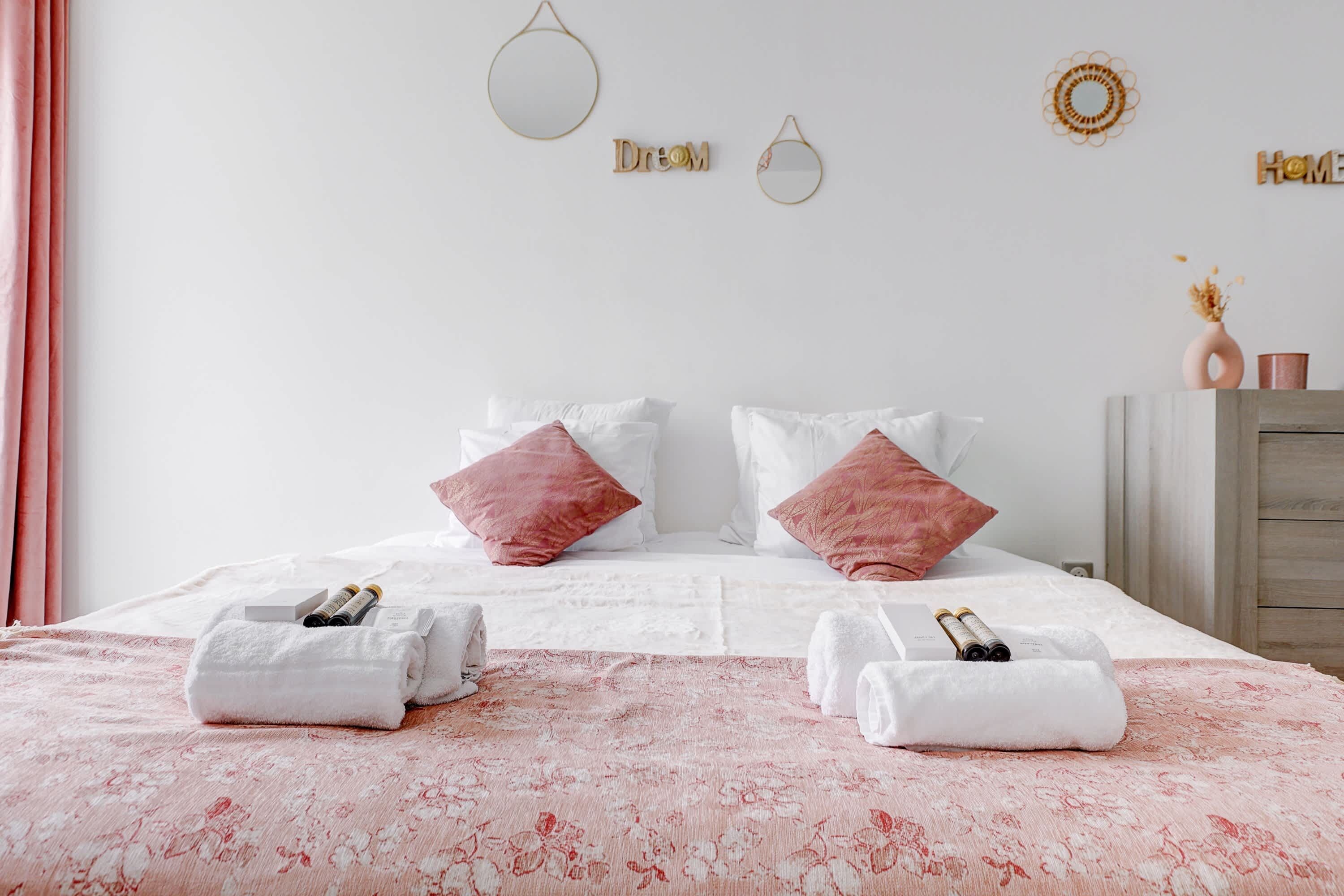 A neatly made bed with white and pink pillows, a floral bedspread, and two towels with toiletries.