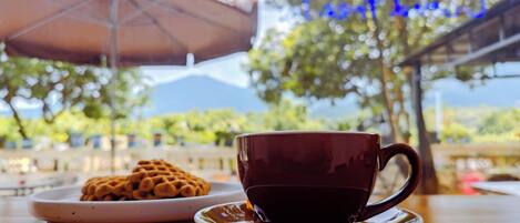 A cup of coffee on a saucer with a plate of waffles in the foreground, an umbrella, and a blurred outdoor setting with trees and a mountain in the background.
