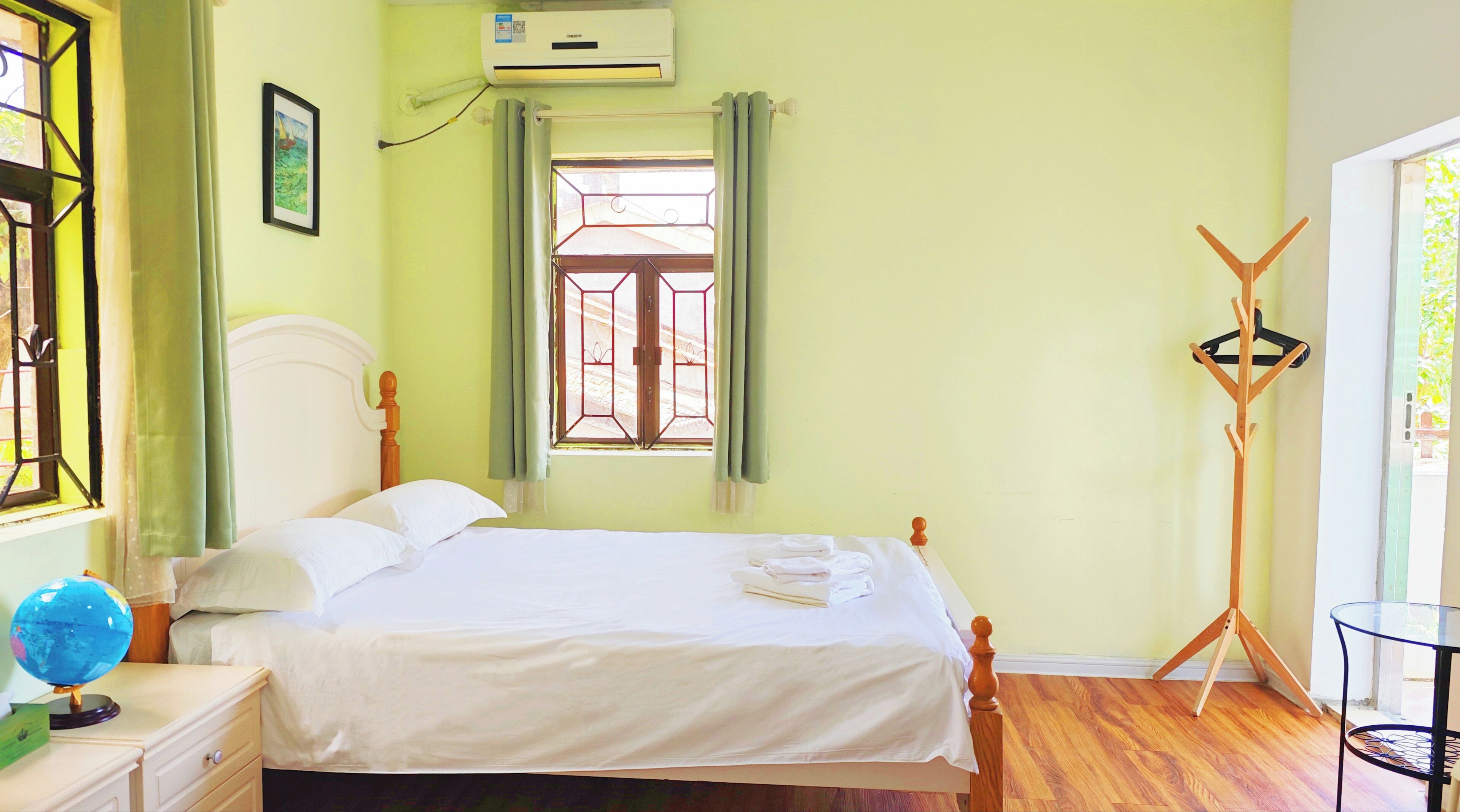 Ein Schlafzimmer mit Bett, Nachttisch mit Globus, Fenster mit Vorhängen, Klimaanlage und Kleiderständer.
