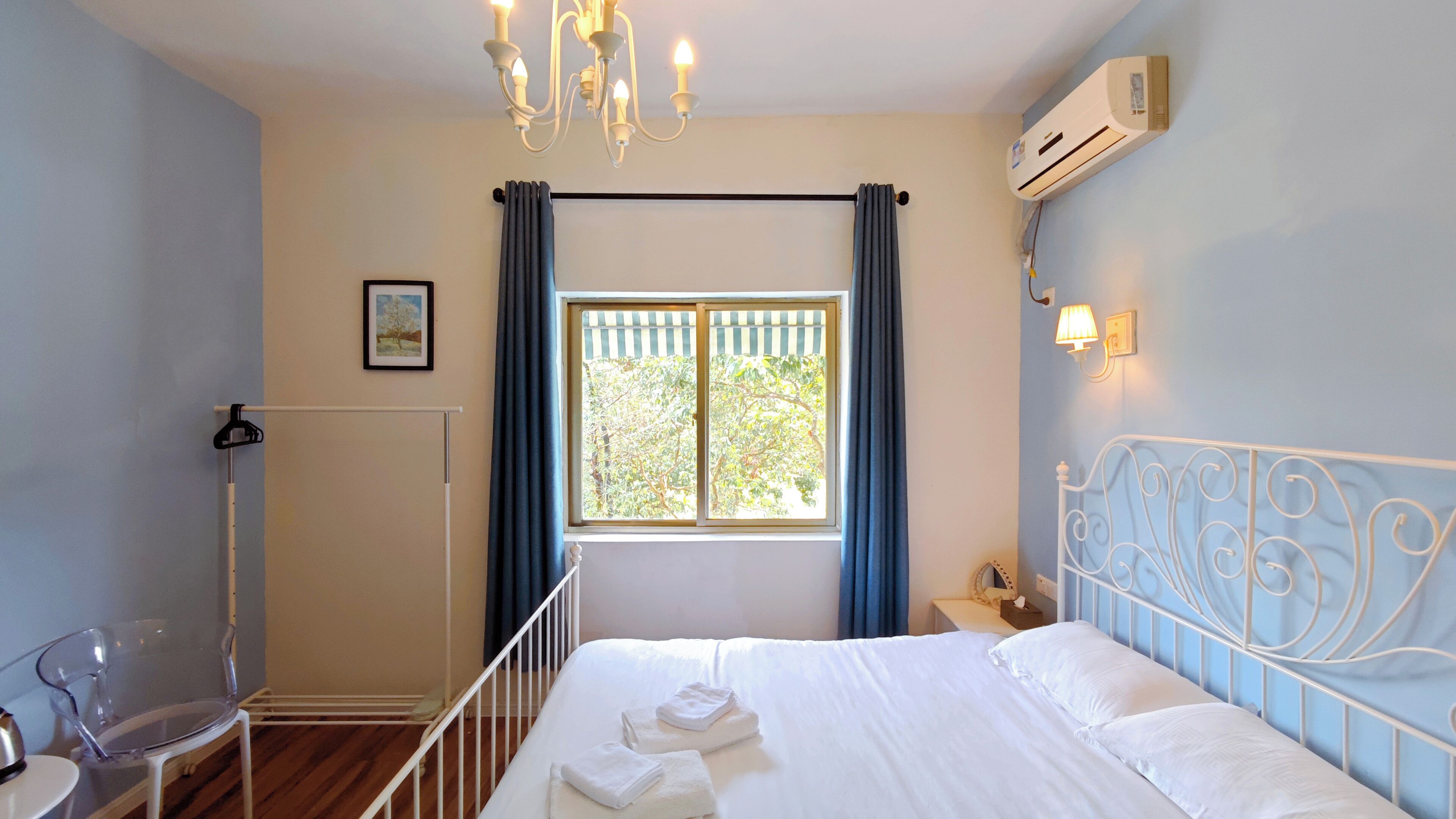 A bedroom with a bed, a chair, a radiator, a window with curtains, a wall-mounted air conditioner, and a chandelier.