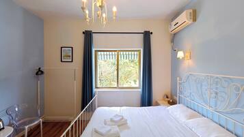 A bedroom with a bed, a chair, a radiator, a window with curtains, a wall-mounted air conditioner, and a chandelier.