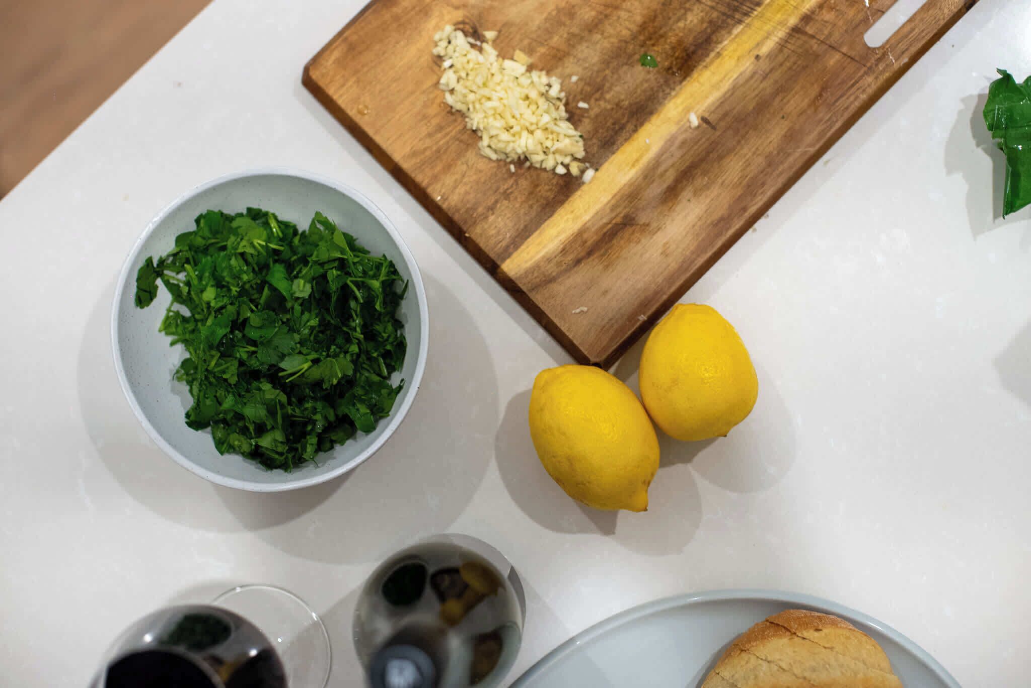 Una encimera de cocina con una tabla de cortar, un bol de hierbas picadas, dos limones y un plato con un pastel.