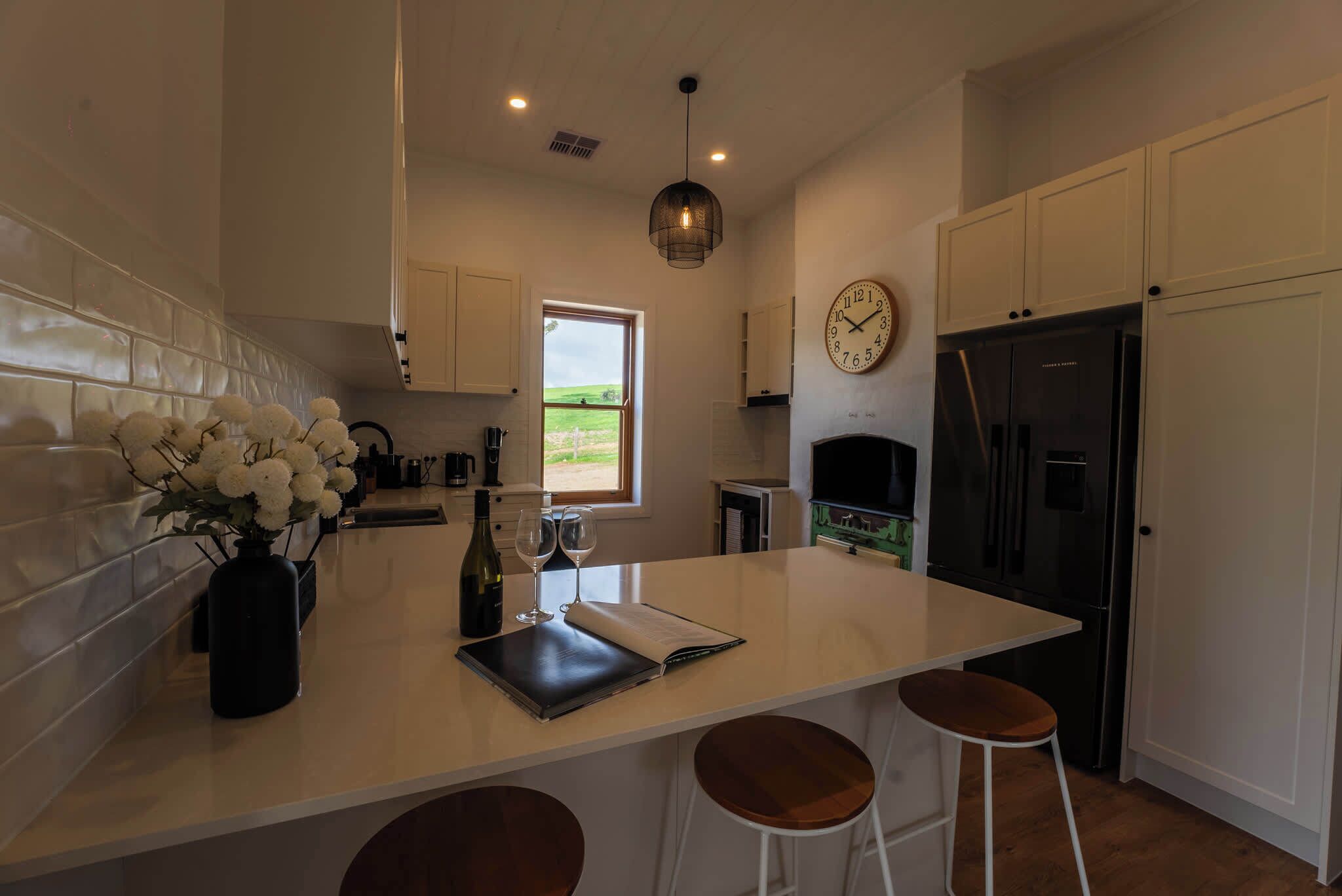 Une cuisine moderne dotée d’un îlot central, de meubles blancs, d’un réfrigérateur noir et d’une fenêtre donnant sur un paysage verdoyant.
