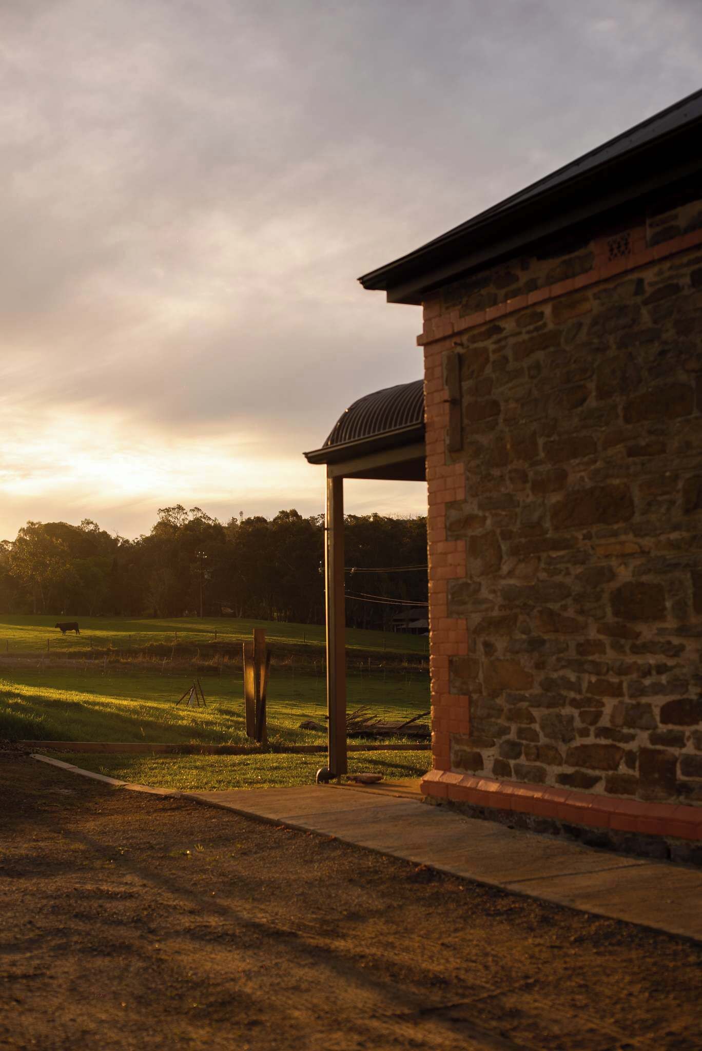 A stone building with a covered outdoor area, a grassy field, and trees in the background.