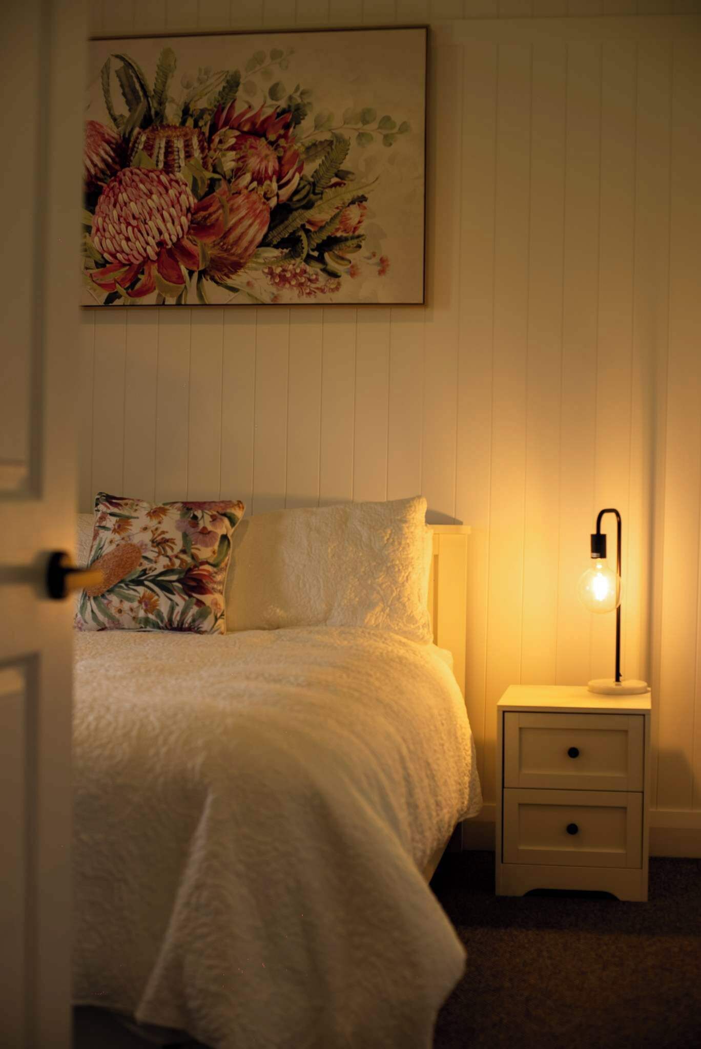 Un dormitorio con cama, una mesita de noche con lámpara, un cuadro floral en la pared y una puerta entreabierta.