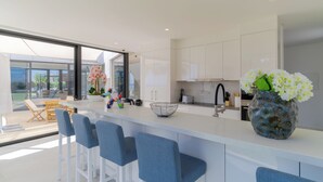 A modern kitchen with white cabinets, a large island with bar stools, a vase of white flowers, and a view of the outdoors. - Luxury Diamond House (Funchal)