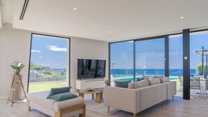 A modern living room with a large sofa, a coffee table, and a TV. There is a large window offering a view of the ocean and a pool. - Luxury Diamond House (Funchal)
