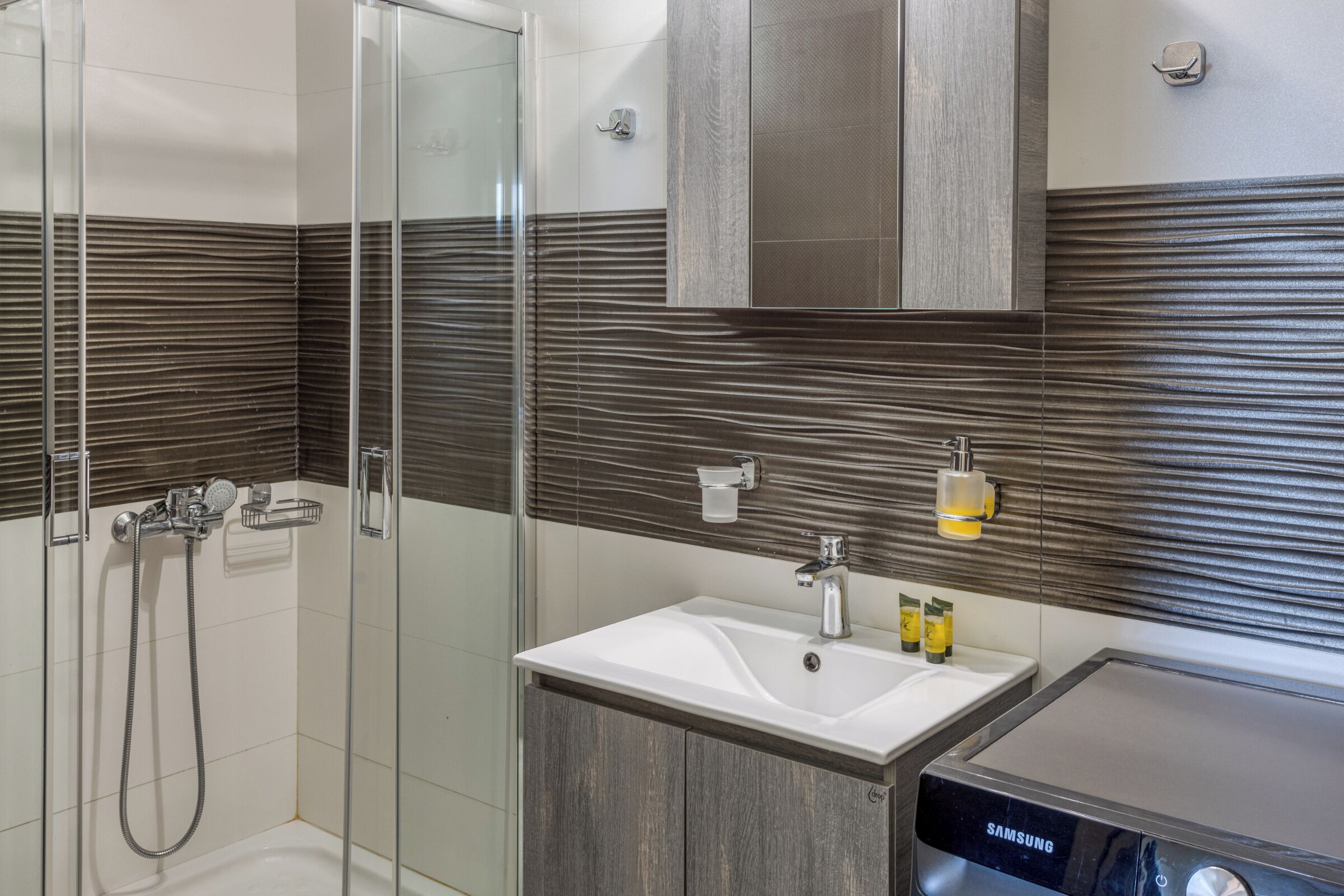 A modern bathroom with a glass shower enclosure, a white sink with a Samsung device, and wooden paneling on the walls.
