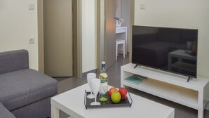 Moderni olohuone, jossa on harmaa sohva, valkoinen sohvapöytä, jonka päällä on hedelmävati, pullo ja lasit. Seinälle on asennettu litteänäyttöinen televisio, joka on sijoitettu valkoiselle jalustalle.