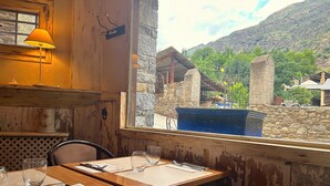 Un comedor rústico con paredes de madera, una ventana que da a un edificio de piedra y vegetación, y una mesa para dos con una vela y una lámpara.