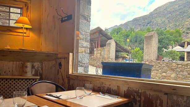 A rustic dining area with wooden walls, a window overlooking a stone building and greenery, and a table set for two with a candle and a lamp. - La Posada d'Àneu by RdA Hotels (Esterri D'Aneu)
