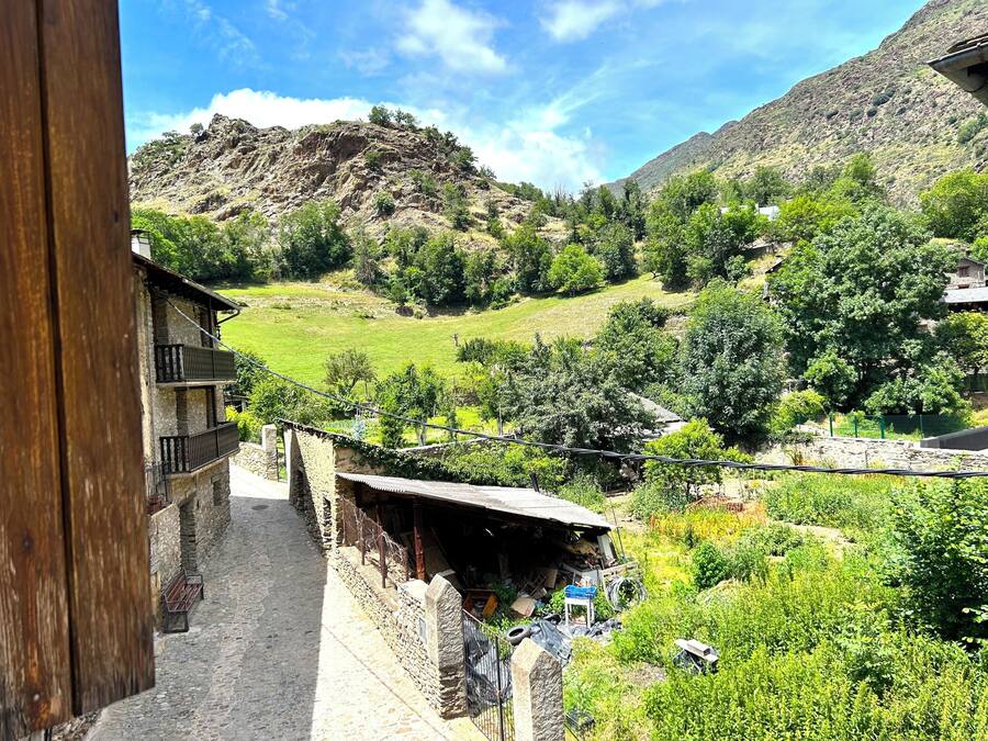 Un edificio rústico de piedra con un balcón, un muro de piedra y vistas a un valle verde con árboles y una montaña al fondo.