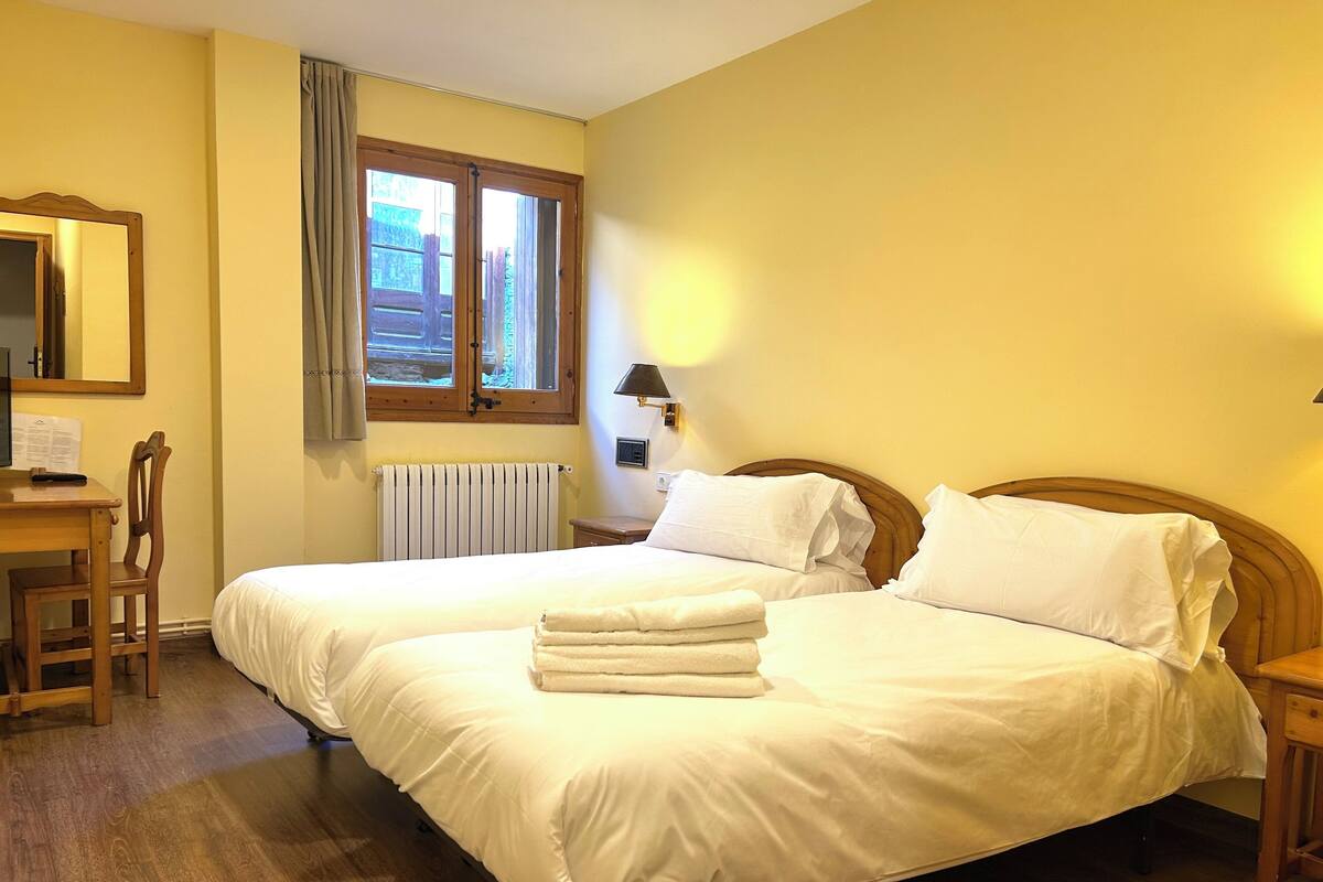 Une chambre d’hôtel avec deux lits, un bureau en bois, une chaise, une fenêtre avec des rideaux, un radiateur et une lampe fixée au mur.