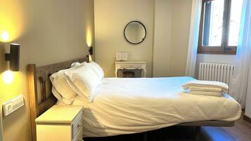 A neatly made bed with white linens, a bedside table with a lamp, a small mirror, and a window with curtains.