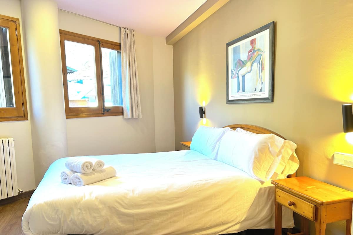 Un lit bien fait, avec du linge de lit blanc et des serviettes pliées, une table de chevet avec une lampe et un tableau accroché au mur.