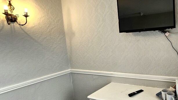 A room with a white wall, a mounted TV, a small white table with a remote control, and a wall-mounted light fixture. - Sapphire Hotel Blackpool (Blackpool)