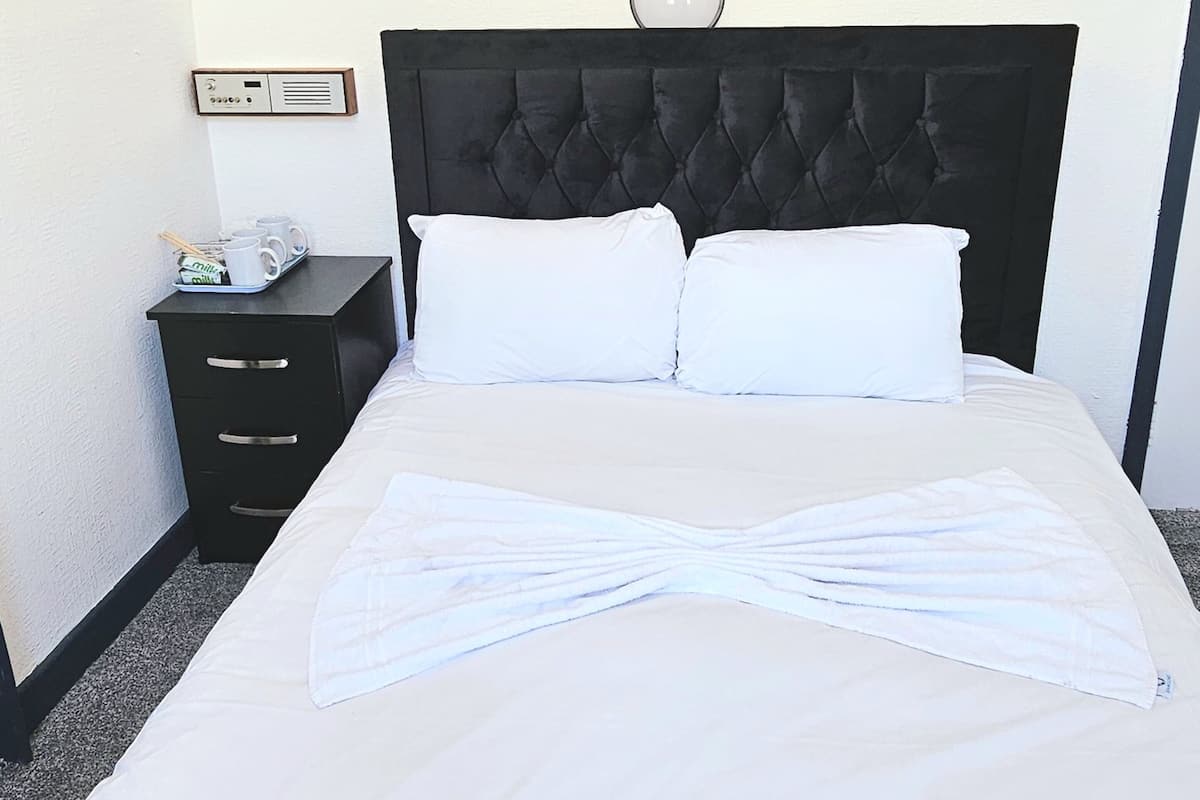 Una cama bien hecha con ropa blanca y una toalla doblada en el centro. Hay una mesita de noche negra con un reloj pequeño y un porta vasos con tazas y una taza.