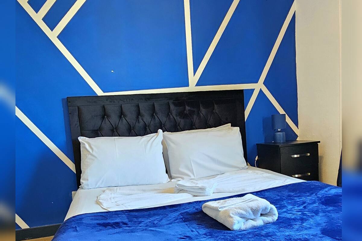Una cama con cabecera azul y ropa de cama blanca, una mesita de noche con lámpara y un diseño geométrico en la pared.