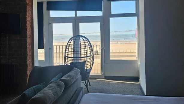 A hotel room with a bed, a seating area with cushions, a chair, and a view of the beach through large windows. - Sapphire Hotel Blackpool (Blackpool)