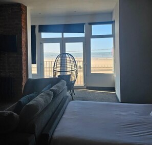 Ein Hotelzimmer mit einem Bett, einer Sitzecke mit Kissen, einem Sessel und Blick auf den Strand durch große Fenster.